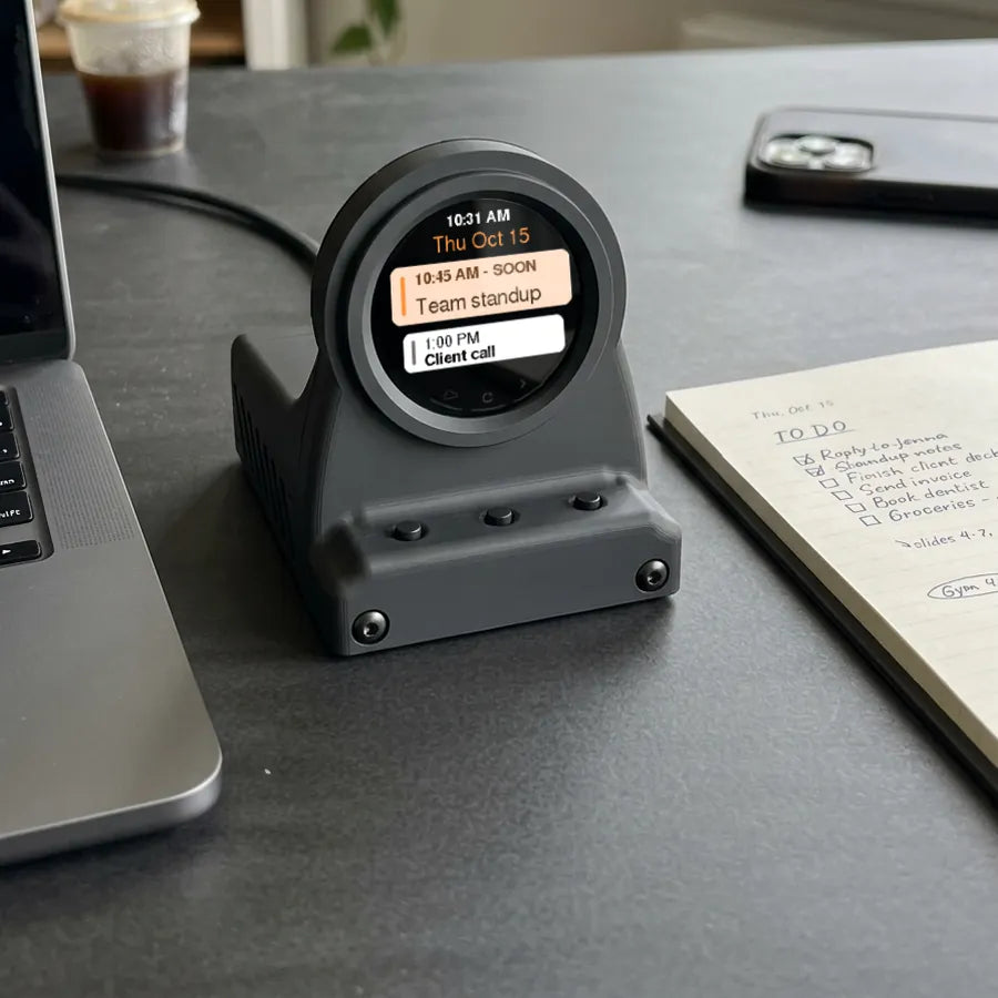 Charcoal Black DeskOrbie 2 on a desk beside a laptop, showing a team standup coming up at 10:45 AM